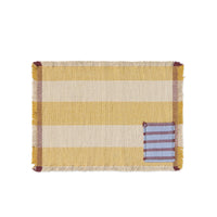 A rectangular woven placemat with horizontal stripes in mustard yellow, cream, and mauve, finished with fringed edges and a small blue striped patch on the bottom right, shown on a plain white background.