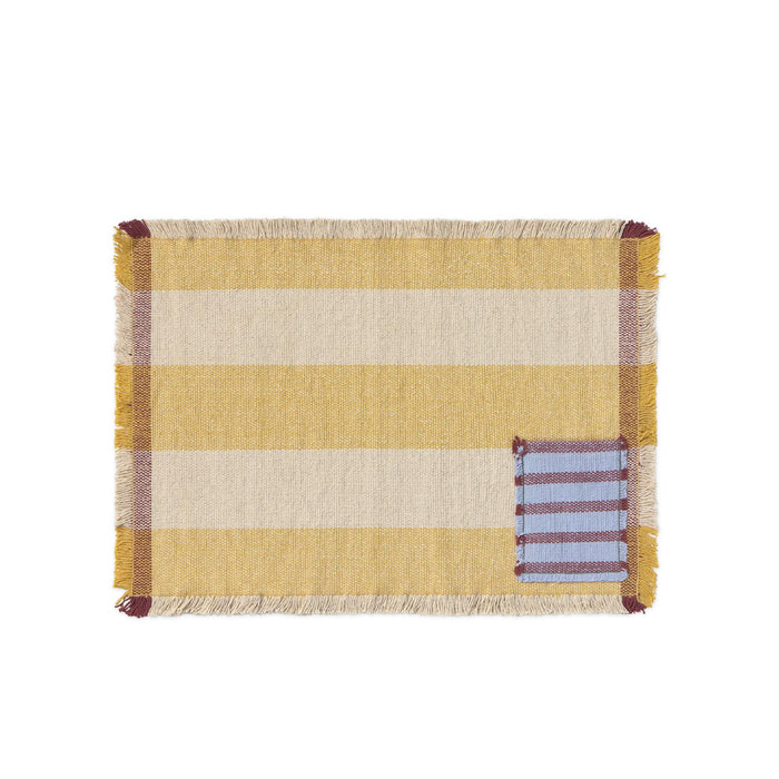 A rectangular woven placemat with horizontal stripes in mustard yellow, cream, and mauve, finished with fringed edges and a small blue striped patch on the bottom right, shown on a plain white background.