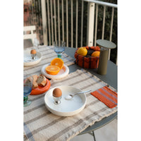 A styled outdoor dining scene on a round table with a gray metal railing. The table is set with a beige-and-gray striped placemat, a white ceramic egg cup holder, an orange egg, a spoon, sliced oranges, bread, and a small woven basket of fruit.