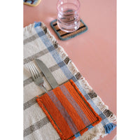 A woven table setting featuring a plaid placemat in beige, blue, and gray with fringe, a matching orange striped napkin, silver cutlery, and a small glass of water on a pink tabletop.