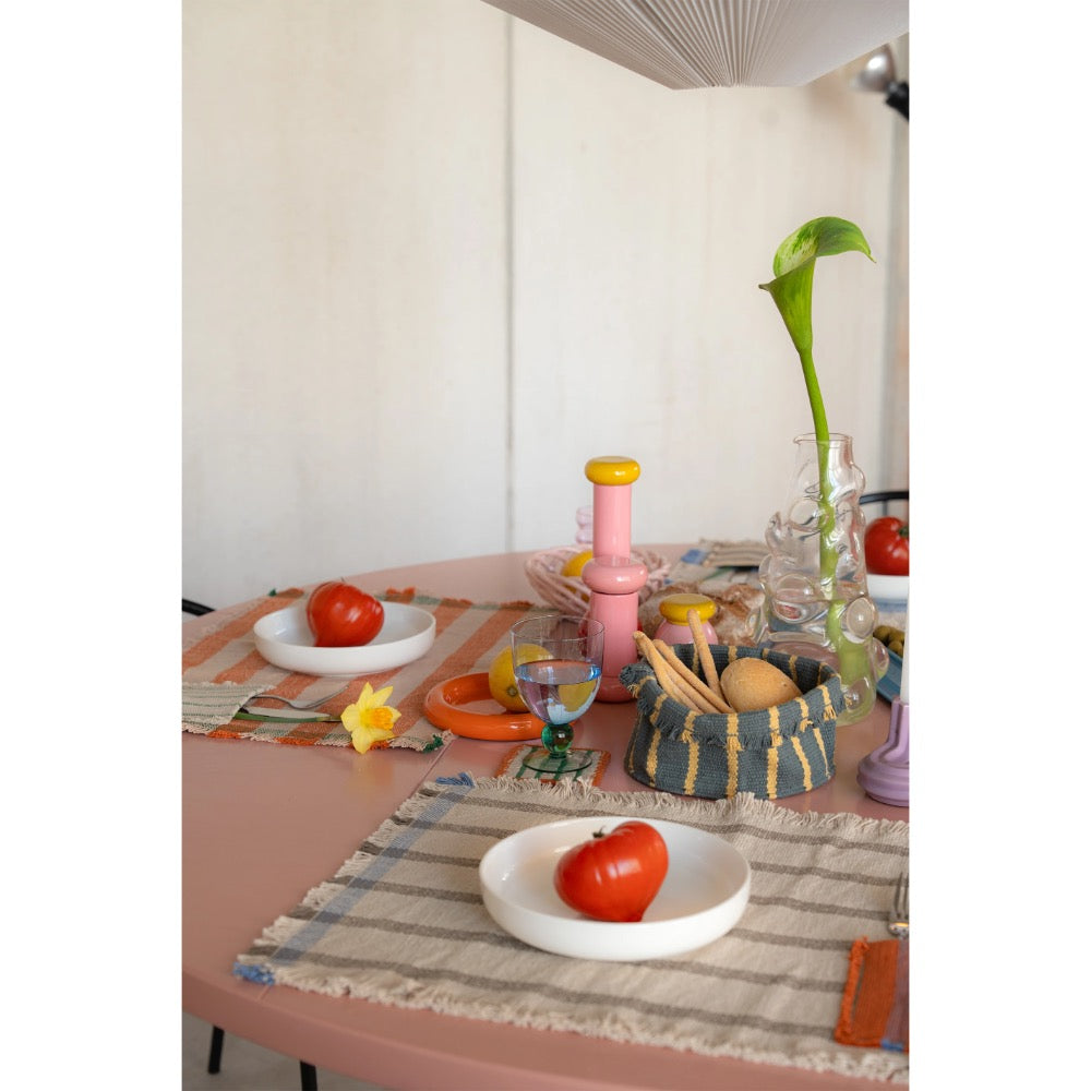 A styled dining table setting with a pink table, striped placemats, white plates holding red tomatoes, a pink bottle, a woven basket, and a glass vase with a green calla lily against a white wall.