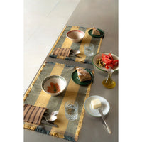 Two woven table runners with fringed edges in a mustard yellow and slate blue striped pattern, styled on a gray dining table with plates, cutlery, and food.