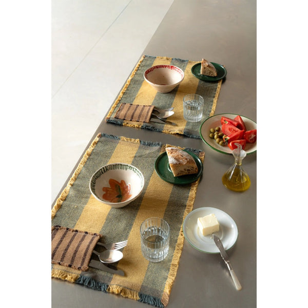 Two woven table runners with fringed edges in a mustard yellow and slate blue striped pattern, styled on a gray dining table with plates, cutlery, and food.