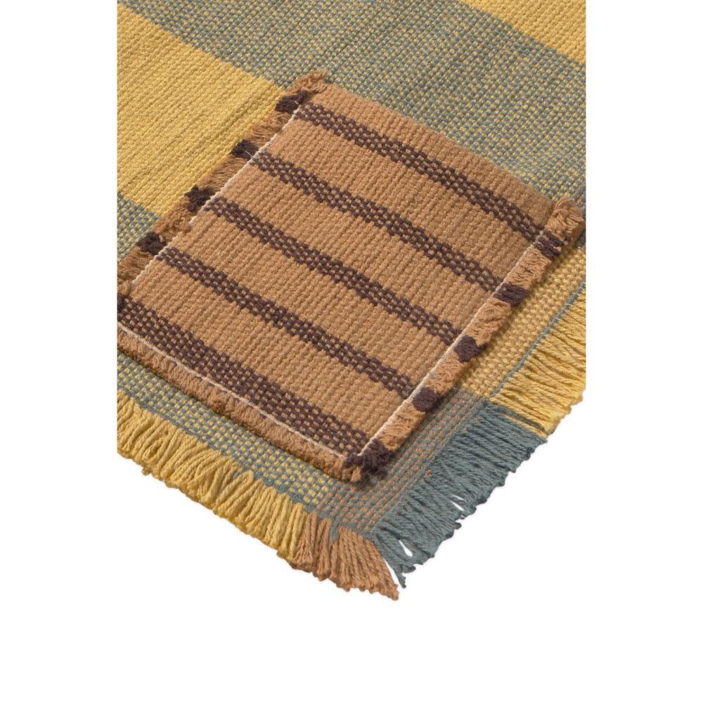 A close-up of a woven placemat showing a mustard yellow and muted blue-green plaid pattern with a central square patch of brown and tan stripes and fringed edges.