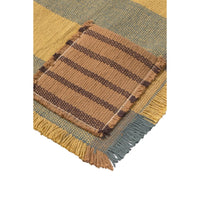 A close-up of a woven placemat showing a mustard yellow and muted blue-green plaid pattern with a central square patch of brown and tan stripes and fringed edges.
