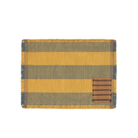 A rectangular woven placemat with horizontal stripes in mustard yellow and dark gray, finished with fringed edges and a small brown striped patch on the lower right, shown on a plain white background.