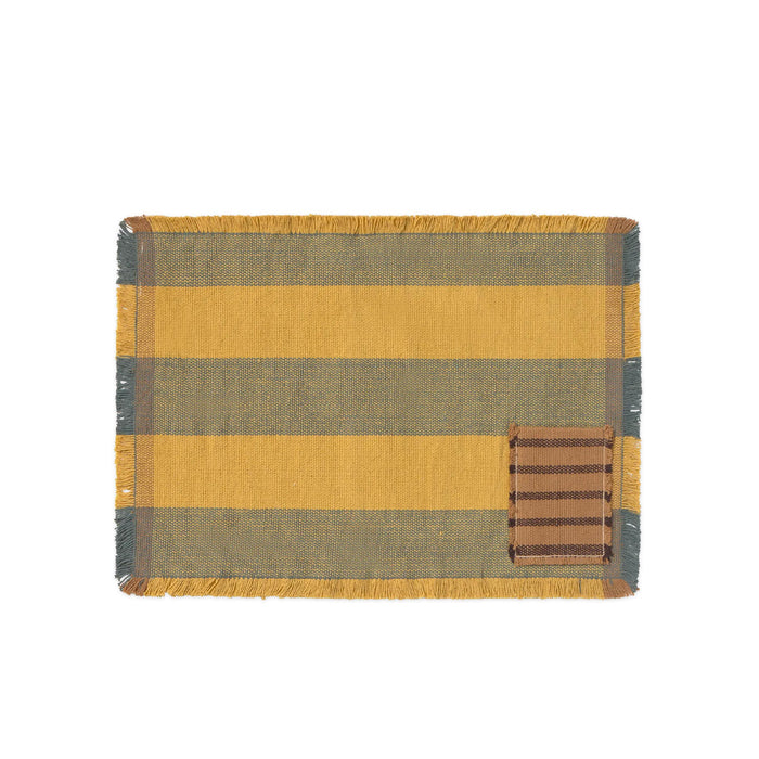 A rectangular woven placemat with horizontal stripes in mustard yellow and dark gray, finished with fringed edges and a small brown striped patch on the lower right, shown on a plain white background.