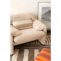 A cream-colored upholstered armchair in a styled living room setting, featuring a striped beige and cream throw pillow, a striped rug, and a framed art print on the wall.
