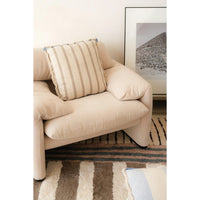 A cream-colored upholstered armchair in a styled room setting, featuring a striped beige and cream throw pillow on the seat. A patterned rug and a framed landscape print are visible in the background.