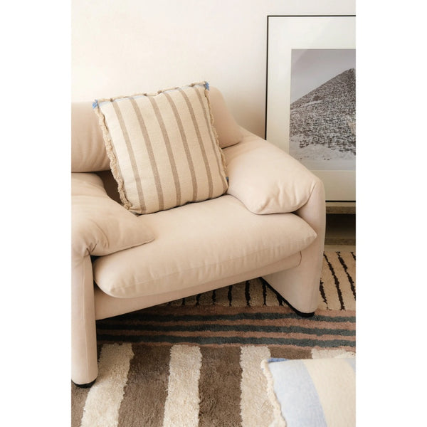A cream-colored upholstered armchair in a styled room setting, featuring a striped beige and cream throw pillow on the seat. A patterned rug and a framed landscape print are visible in the background.