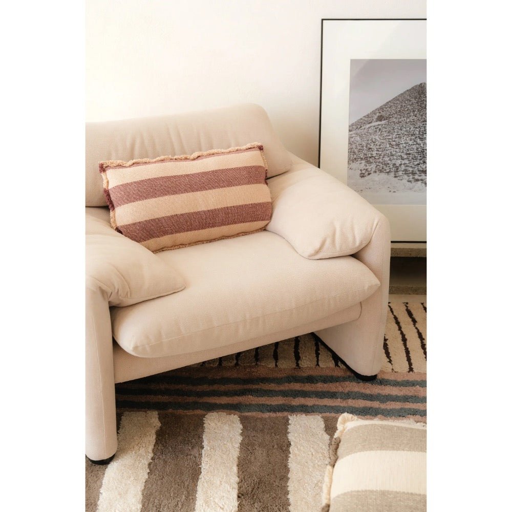 A cream-colored upholstered armchair in a styled room setting, featuring a striped throw pillow in beige and rust tones, with a striped rug and a framed art print in the background.