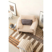 A beige upholstered armchair in a styled room setting, featuring a dark gray striped throw pillow. The chair is placed on a brown and beige striped area rug, with a matching striped throw pillow in the foreground and framed wall art on the walls.