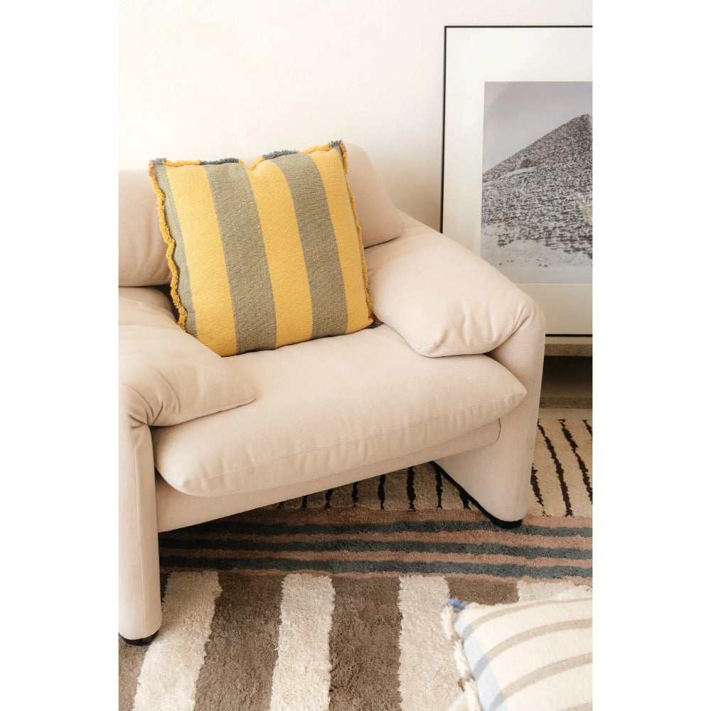 A cream-colored armchair in a styled living room setting, featuring a striped throw pillow in yellow and gray on the seat. A striped rug and a framed landscape print are visible in the background.