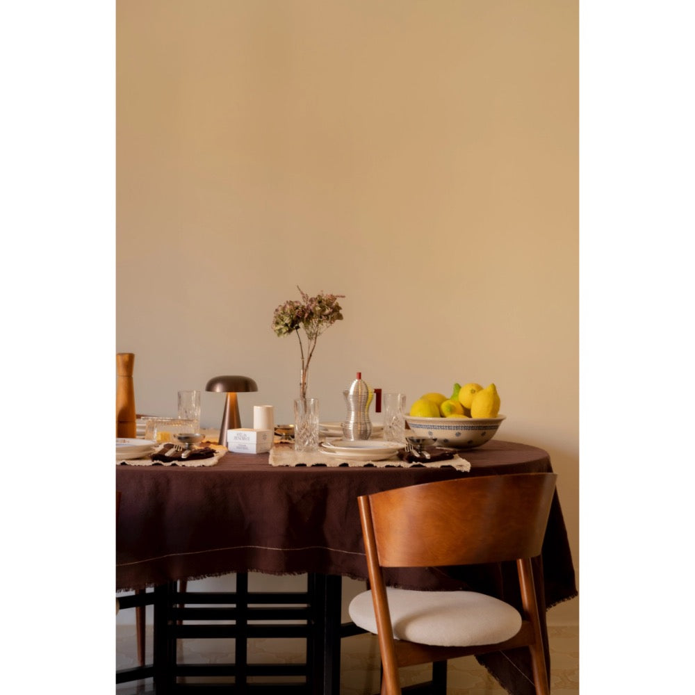 A styled dining table in a warm neutral room, featuring a dark brown tablecloth, a wooden chair with a cream cushion, a bowl of yellow pears, and a small floral arrangement.