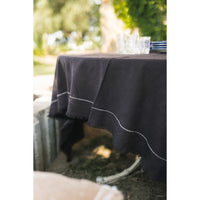 A dark brown tablecloth with white contrast stitching and frayed edges, draped over an outdoor table with a glass and plates on top, set against a blurred garden background.
