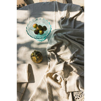 A draped light beige tablecloth, styled on a wooden surface with a glass bowl of olives and a small decorative object under warm, dappled sunligght.