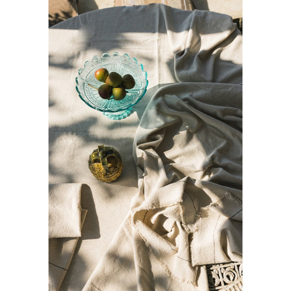 A draped light beige tablecloth, styled on a wooden surface with a glass bowl of olives and a small decorative object under warm, dappled sunligght.