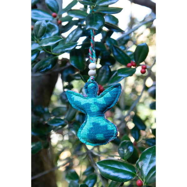 A teal green angel-shaped fabric ornament hanging from a string with white beads, set against a background of green holly leaves and small red berries.
