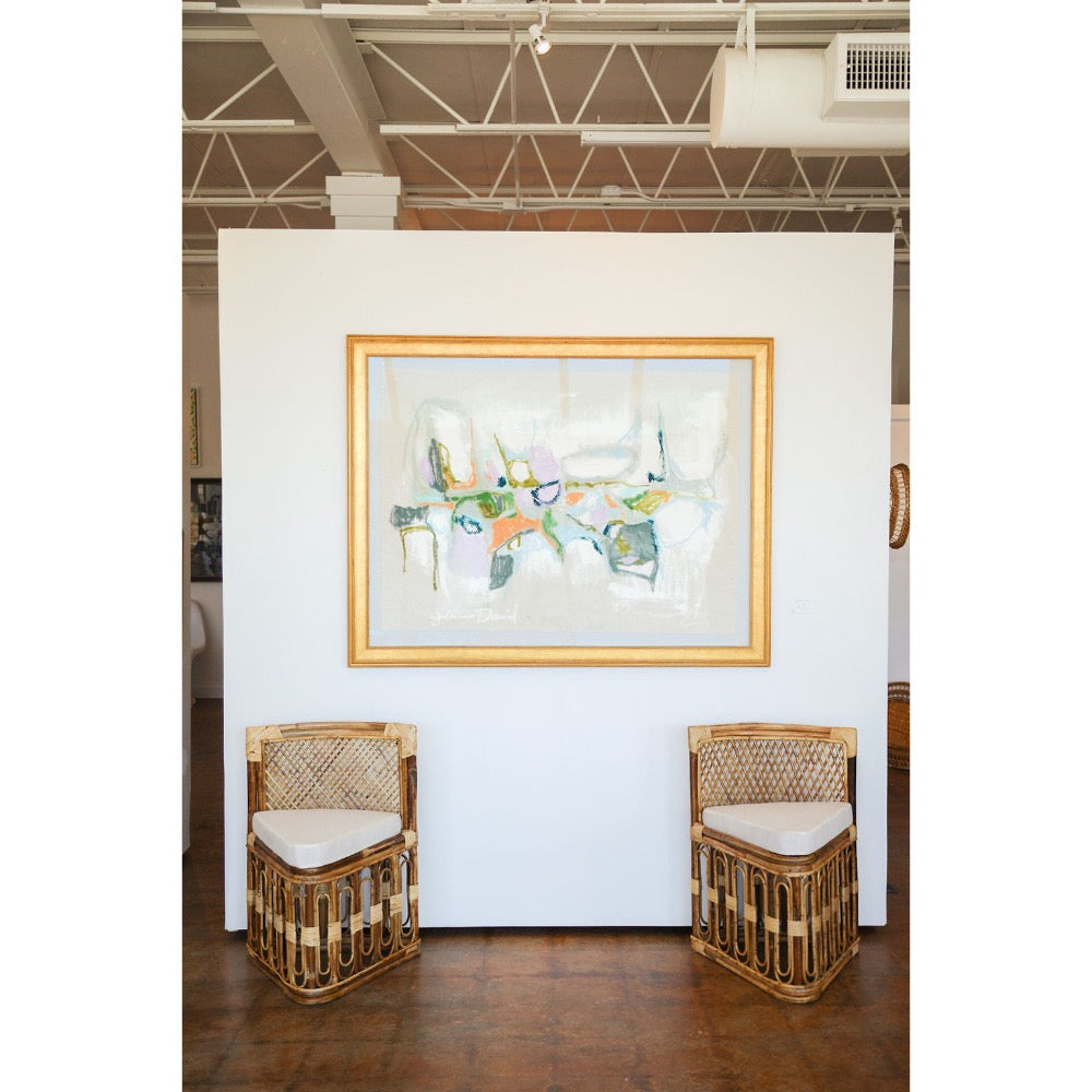 Two wicker chairs with white cushions in front of a colorful abstract painting on a white wall.