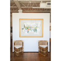 Two wicker chairs with white cushions in front of a colorful abstract painting on a white wall.