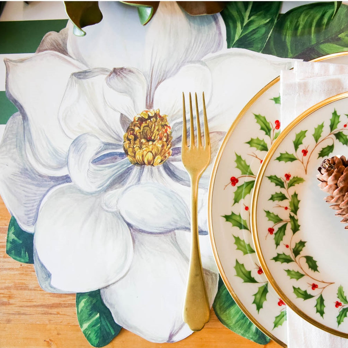 A gold fork on a Magnolia Die Cut Paper Placemat by Hester & Cook beside holiday plates and a pinecone napkin.