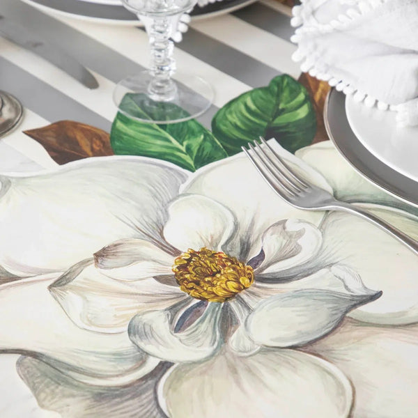 A Magnolia Die Cut Paper Placemat by Hester & Cook on a striped table set with a fork, plate, and glass.