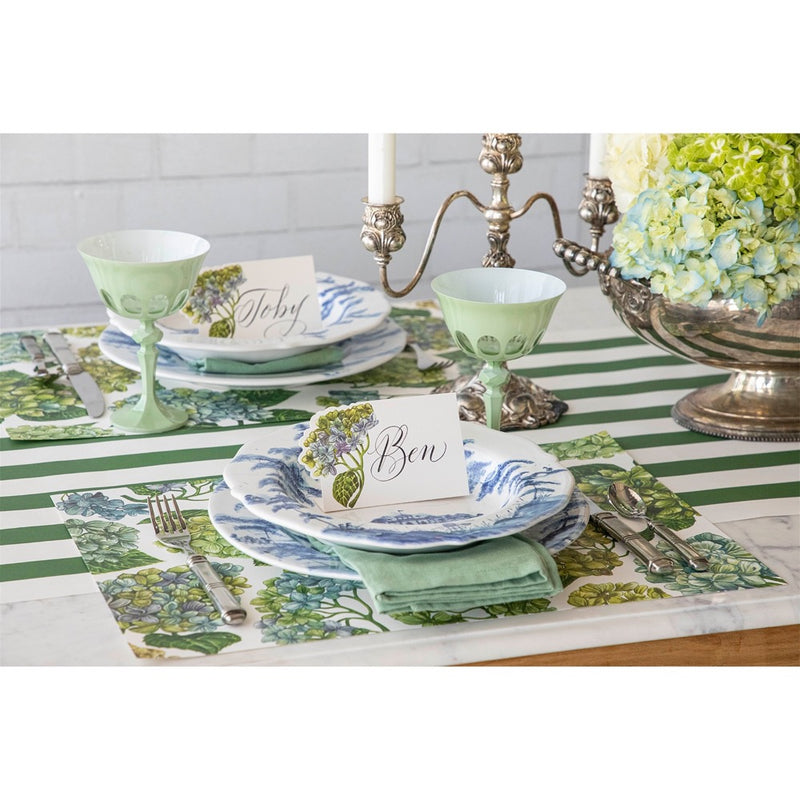 Elegant table setting featuring floral plates, green goblets, candles, and name cards reading "Seb" and "Ben" on a Hester & Cook Dark Green Classic Stripe Paper Runner.