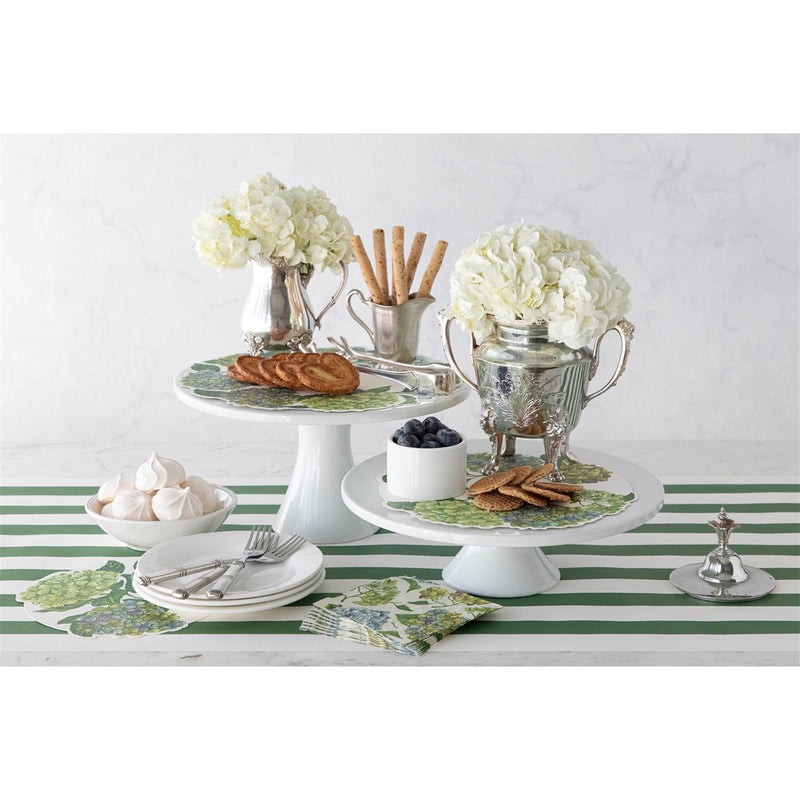 A table set with flowers, pastries, and decorative items on white cake stands and a Dark Green Classic Stripe Paper Runner by Hester & Cook.