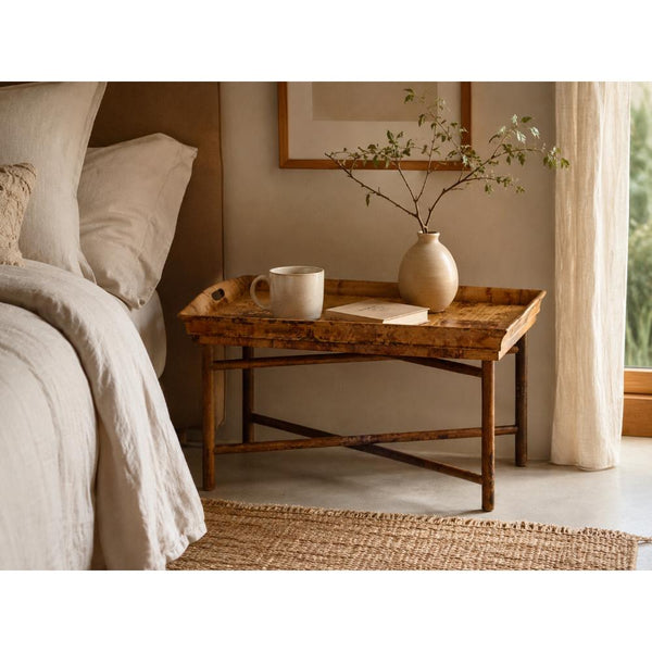 A rustic wooden nightstand tray styled in a bedroom next to a bed, holding a white ceramic mug, a small beige vase with branches, and a book.