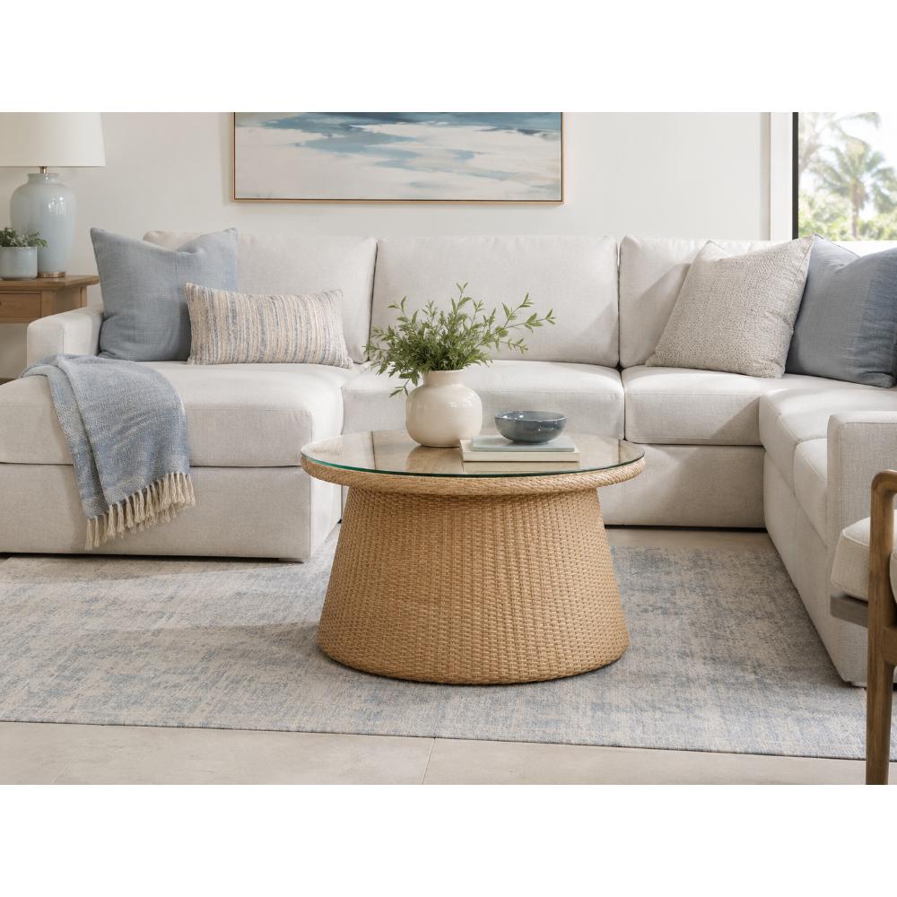 A round woven coffee table with a glass top sits in front of a light beige sectional sofa styled with neutral throw pillows and a blue blanket in a bright, modern living room.