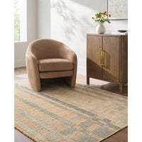A modern taupe armchair sits on a striped beige and gray area rug in a bright, contemporary room next to a wooden cabinet with gold handles.