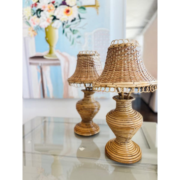 Two wicker table lamps on a reflective surface with a blurred floral arrangement in the background.