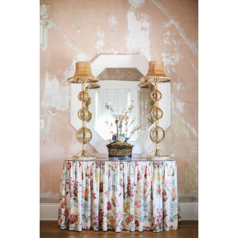 Decorative table with floral skirt and wicker lamps against a pink wall.