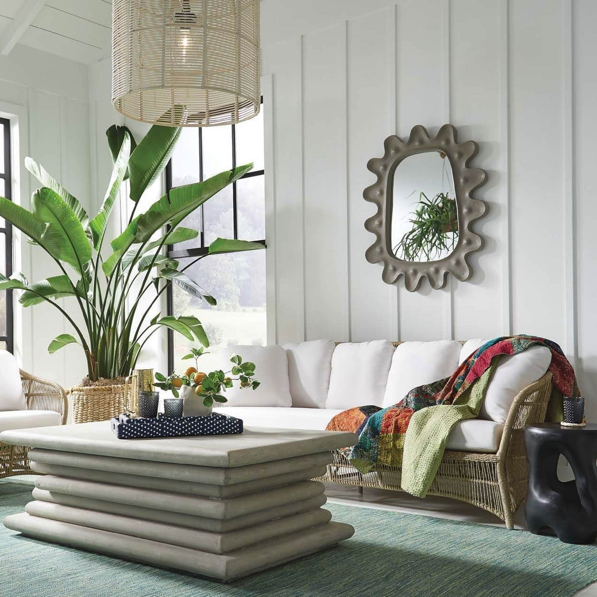 The bright living room features a white couch and a large plant, with the Lukas Mirror by Made Goods adding modern flair with its sleek gray-colored concrete design. A layered coffee table rests elegantly on a blue-green rug, creating a harmonious entryway decor.