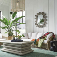 The bright living room features a white couch and a large plant, with the Lukas Mirror by Made Goods adding modern flair with its sleek gray-colored concrete design. A layered coffee table rests elegantly on a blue-green rug, creating a harmonious entryway decor.