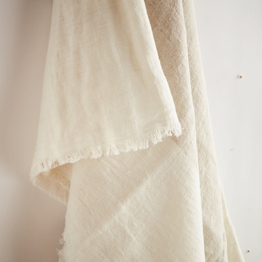 A close-up of a draped off-white or cream textured throw with frayed edges, hanging against a plain light background.