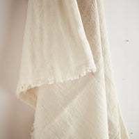 A close-up of a draped off-white or cream textured throw with frayed edges, hanging against a plain light background.
