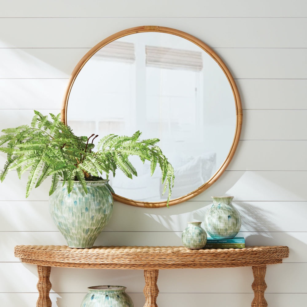 Round mirror with rattan frame above a wicker console table with decorative vases and a fern.