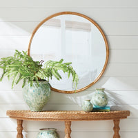 Round mirror with rattan frame above a wicker console table with decorative vases and a fern.