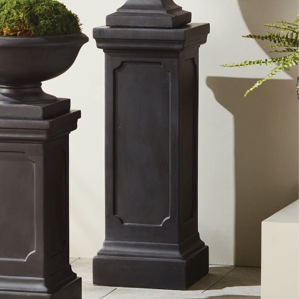 The Fibreclay Chambers Pedestal by Napa Home & Garden features an antique black finish beside a concrete planter and greenery.