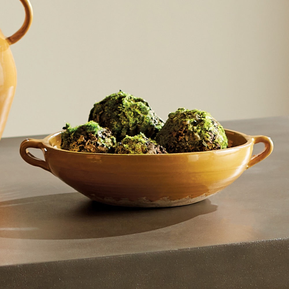 A rustic yellow-brown ceramic bowl with side handles, filled with green moss-topped balls, set on a dark tabletop against a neutral beige background.