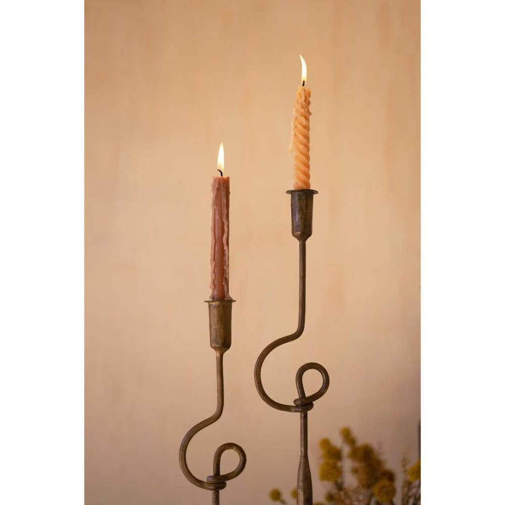 Two lit taper candles in dark metal scroll candlesticks against a warm beige background.