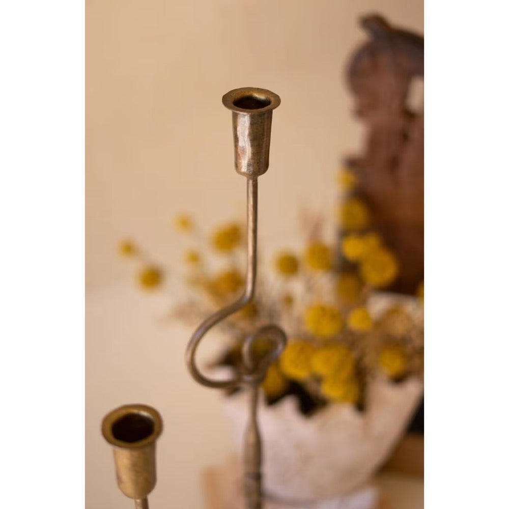 A rustic metal candlestick holder with a curved stem and two cup-like candle sockets, shown in a softly blurred indoor setting with yellow flowers in the background.