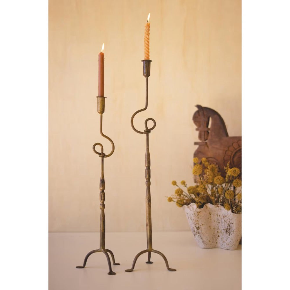 Two tall metal candlestick holders with scrolled arms and tripod bases, each holding a lit taper candle, displayed indoors against a warm beige wall with a decorative horse and flower arrangement in the background.