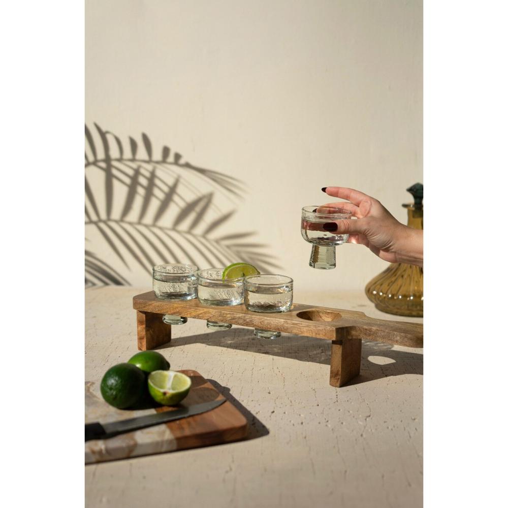Person holding a glass of water next to a wooden stand with small glasses and limes on a light background