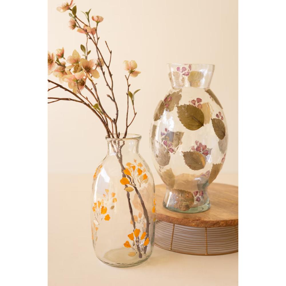 Two clear glass vases decorated with pressed botanical elements, one holding dried flower branches and the other standing alone, set on stacked wood blocks against a warm cream background.