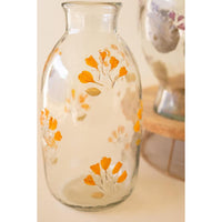 A clear glass bottle vase decorated with hand-painted orange and beige ginkgo leaves, shown on a light beige surface with another blurred glass vase in the background.
