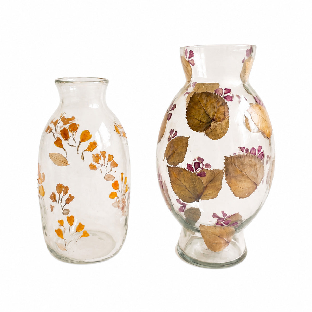 Two clear glass vases decorated with dried botanical elements, including orange and purple flowers and brown leaves, shown on a plain white background.