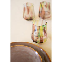 A set of hand-blown glass tumblers with abstract multicolor swirls of green, yellow, brown, and pink on a cream background, with a close-up of matching brown plates in the foreground.