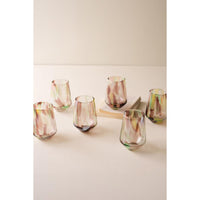 Six small stemless drinking glasses with a marbled, iridescent finish in soft pink, green, and gold tones, arranged on a light beige surface against a neutral cream background.